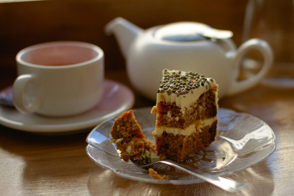 A small white teapot, a cup of tea and a slice of cake on a plate with a fork is seen on a table