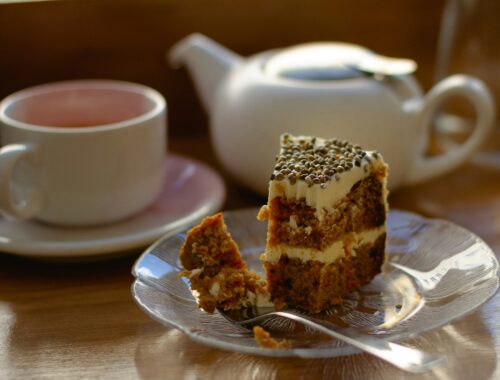 A small white teapot, a cup of tea and a slice of cake on a plate with a fork is seen on a table