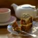 A small white teapot, a cup of tea and a slice of cake on a plate with a fork is seen on a table