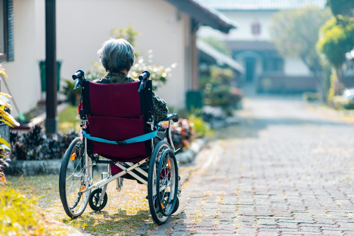 A person seen from behind, is sitting in a wheelchair, alone on a sunny street.