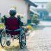 A person seen from behind, is sitting in a wheelchair, alone on a sunny street.