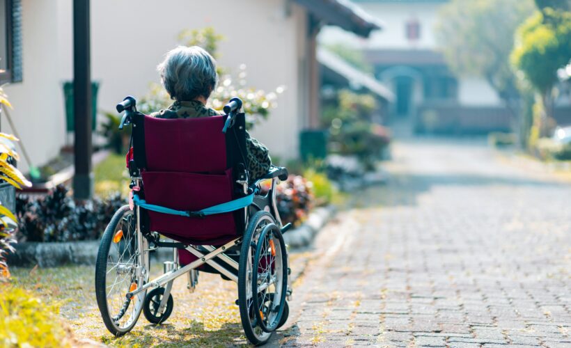 A person seen from behind, is sitting in a wheelchair, alone on a sunny street.