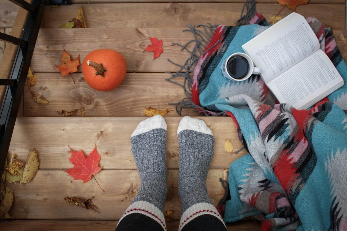 someone's feet in comfy socks is stood on steps with a blanket and a book open beside the feet and pumpkins the other side it looks like they are stood on wooden steps and we are seeing from above