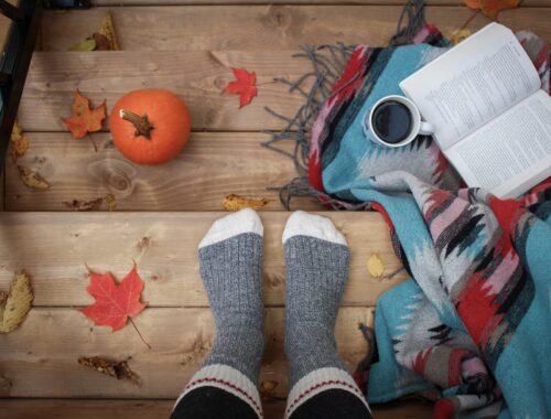 someone's feet in comfy socks is stood on steps with a blanket and a book open beside the feet and pumpkins the other side it looks like they are stood on wooden steps and we are seeing from above