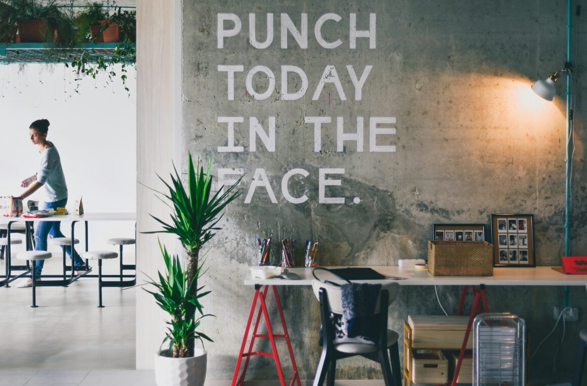 A desk with a green plant beside it is seen, on the wall behind it is written Punch today in the face. A woman is seen beside a dining table in the background