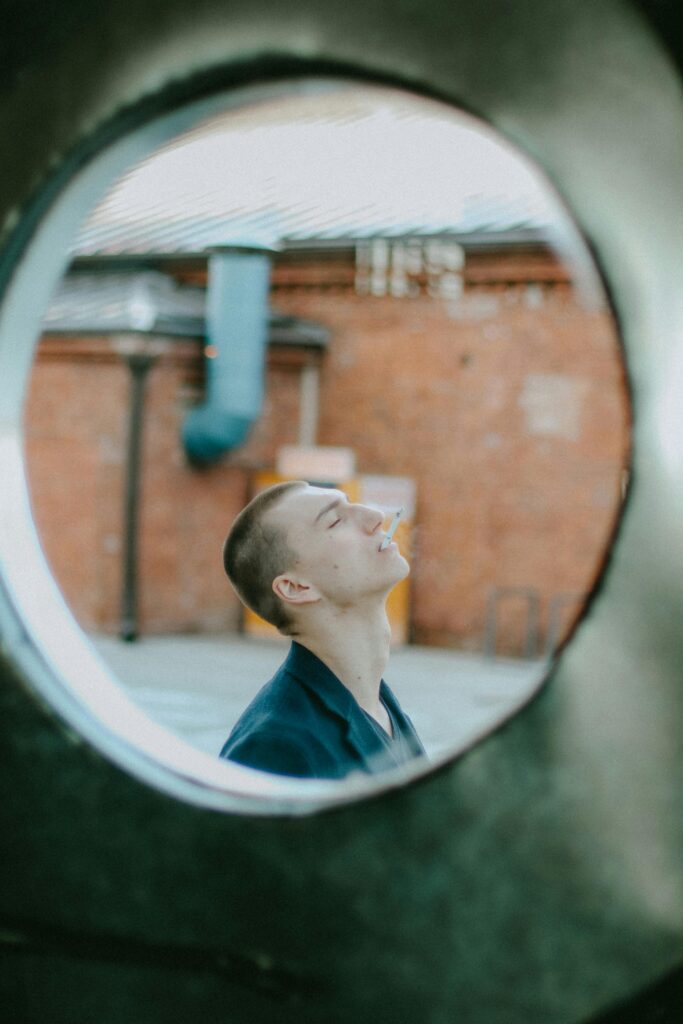 A white man with a shaved head is seen through a circular window looking up and smoking a cigarette 