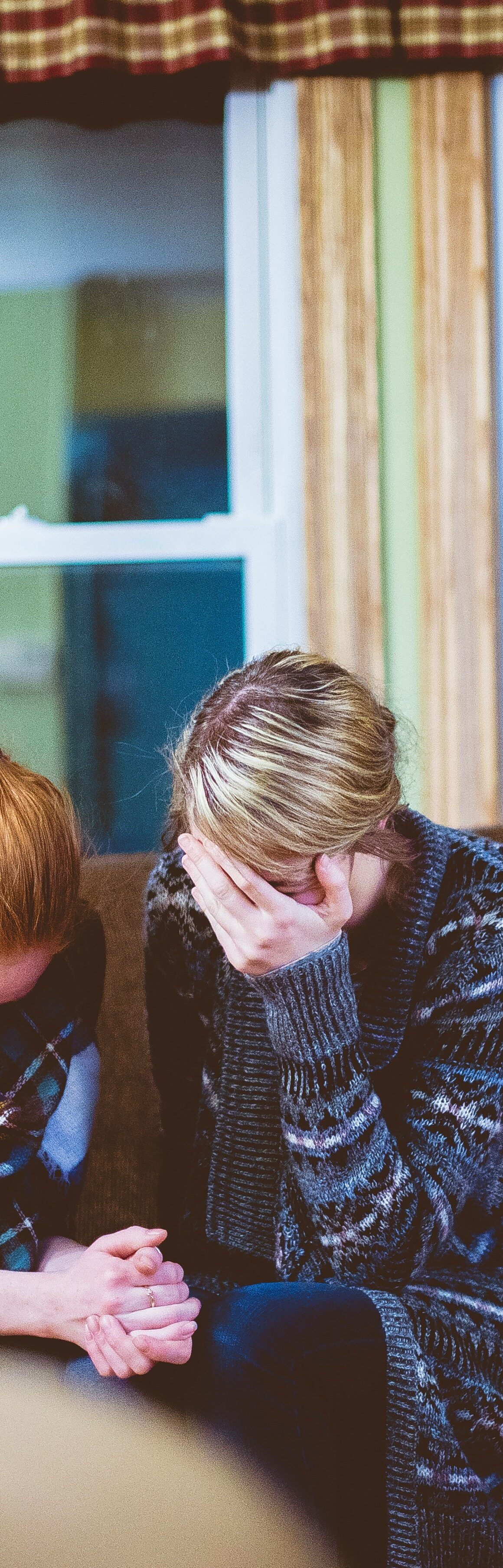 Image description - A blonde woman and a redheaded woman are sat huddled on a brown sofa, there is a large window behind them. They are crying and holding one another