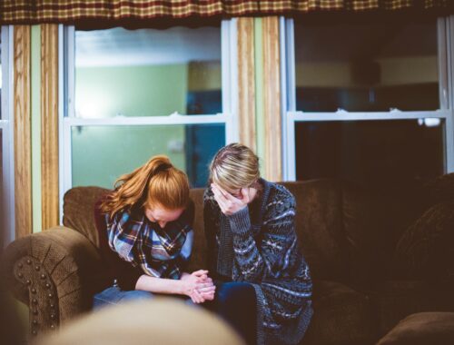 Image description - A blonde woman and a redheaded woman are sat huddled on a brown sofa, there is a large window behind them. They are crying and holding one another