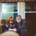 Image description - A blonde woman and a redheaded woman are sat huddled on a brown sofa, there is a large window behind them. They are crying and holding one another