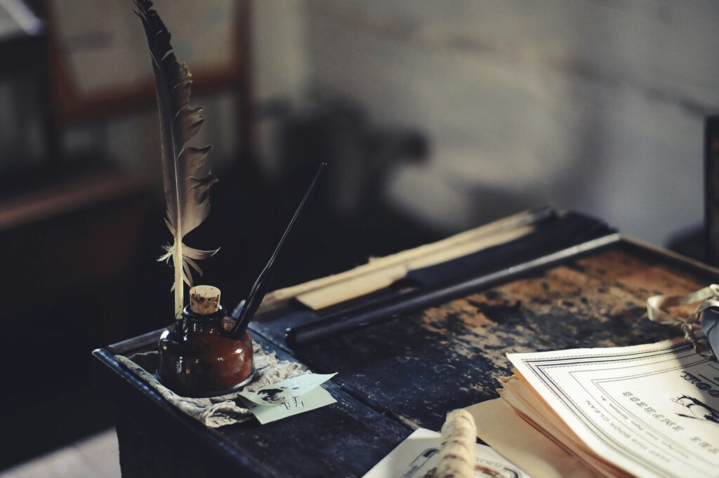 An old desk is seen with a quill standing in an inkwell with some papers on the desk