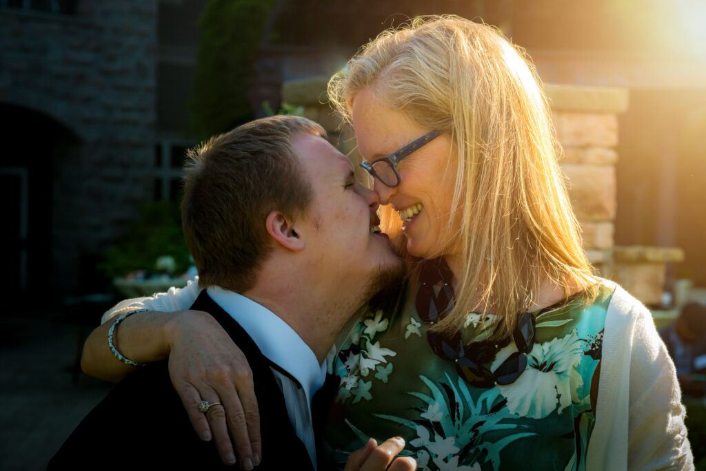 A man with Down Syndrome is smiling while face to face with an older woman wearing glasses and also smiling a sun glare is seen behind them and they are dressed really nice in a suit and blue top 