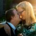 A man with Down Syndrome is smiling while face to face with an older woman wearing glasses and also smiling a sun glare is seen behind them and they are dressed really nice in a suit and blue top