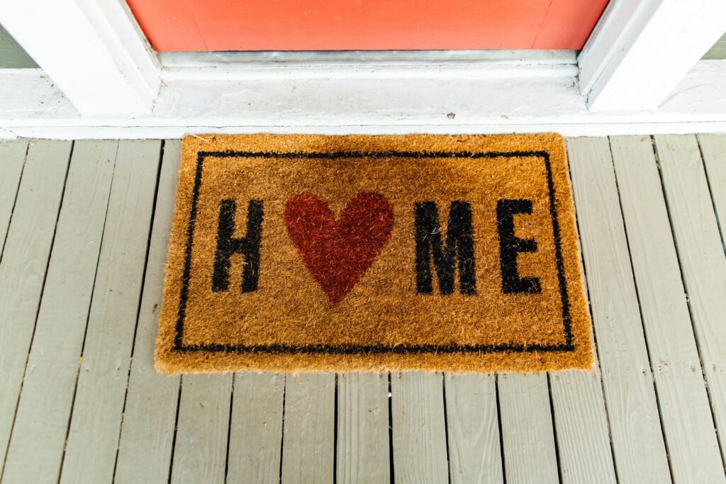 A doormat outside on a white porce with a red door. It says Home where the o is a heart 