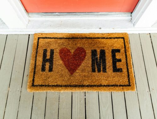 A doormat outside on a white porce with a red door. It says Home where the o is a heart