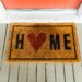 A doormat outside on a white porce with a red door. It says Home where the o is a heart